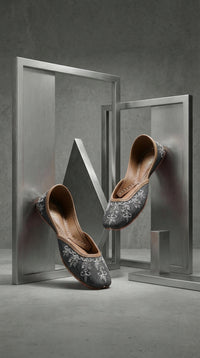 Saanjh grey embroidered jutti pair – artistic display between brushed steel frames by Radiarte