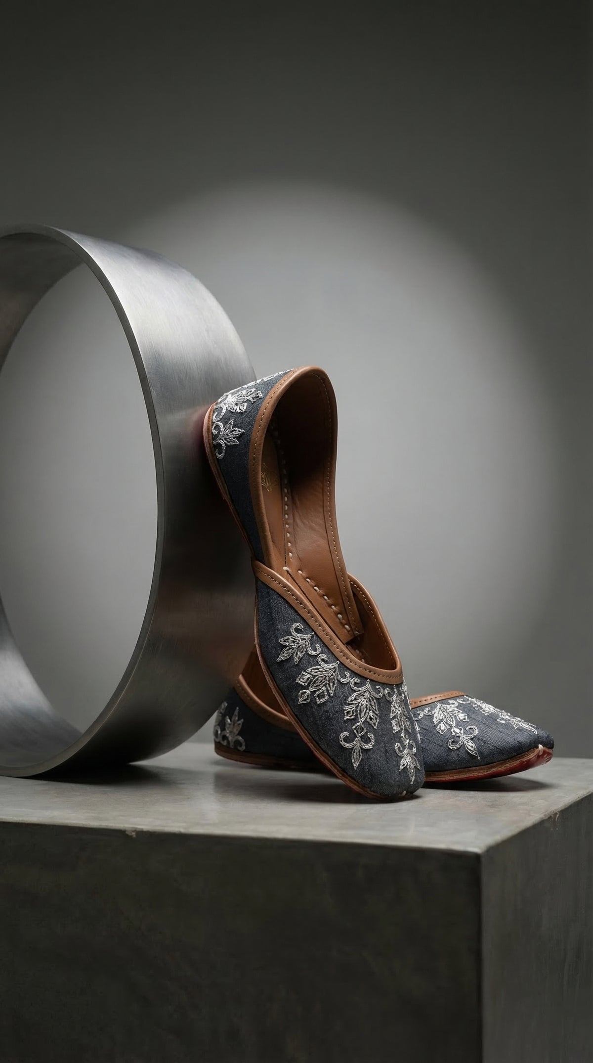 Saanjh grey silk jutti close-up – silver floral and leaf thread embroidery detail against steel cylinder