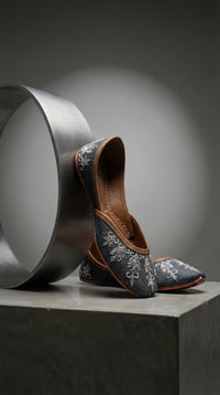 Saanjh grey silk jutti close-up – silver floral and leaf thread embroidery detail against steel cylinder