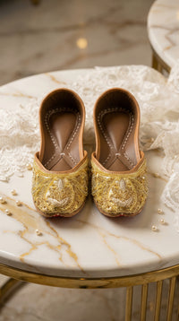 Shararat gold bugle bead jutti pair – front view on marble side table with ivory lace by Radiarte