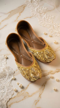 Shararat gold sequin zardozi jutti – top angle flat lay on marble with scattered pearls Radiarte