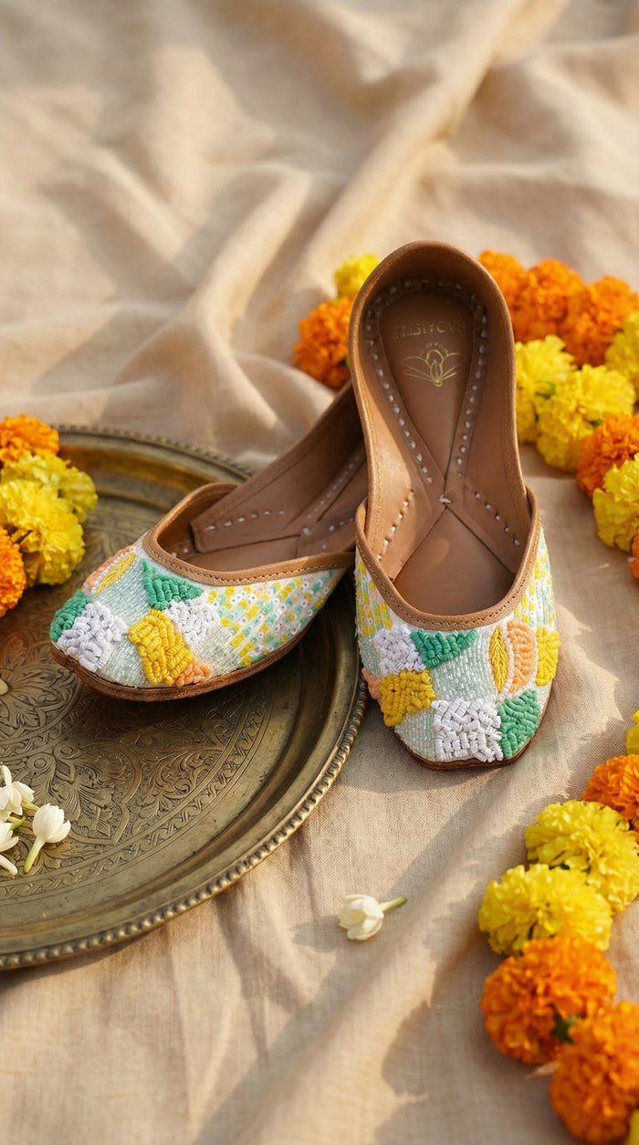 Zale pastel floral jutti pair – display on brass thali surrounded by marigold garland and mogra Radiarte