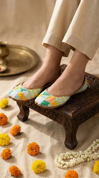 Zale multicolour embroidered jutti on feet – worn with beige linen trousers on carved wooden chowki Radiarte
